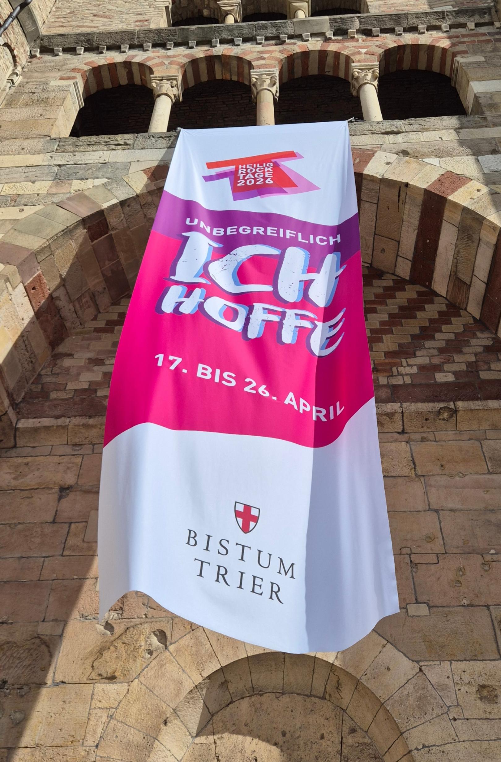 Über dem Eingang zum Dom hängt ein langes Banner im Hochformat. Darauf steht: 'Unbegreiflich: Ich hoffe', die Schrift ist Magenta und Violett unterlegt